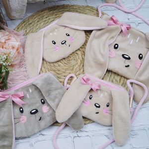 Könnte beinhalten: Drei Plüsch-Handtaschen in Hasenform mit rosa Schleifen und gestickten Gesichtern. Die Taschen sind aus weichem, flauschigem Stoff und haben verstellbare Träger.
