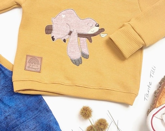 May include: A mustard yellow waffle knit sweatshirt with a brown branch and a pink sloth embroidered on the front. The sweatshirt has a brown patch with a rainbow and the text "Ich bin ein Regenbogen" on the left side of the chest.