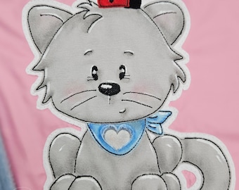 May include: A gray cat with a blue bandana and a red ladybug on its head. The cat is sitting with its paws tucked under its body and its tail curled around its legs. The cat is embroidered on a pink fabric.