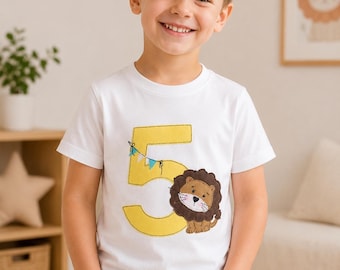 May include: A white short-sleeved t-shirt featuring a large yellow number five appliqué. A small brown lion with a dark brown mane is positioned next to the number, and a string of tiny blue and white triangular pennants hangs from the number five.