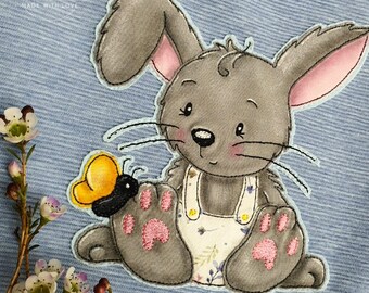 May include: A gray bunny with pink ears and a floral dress sits on a blue background. The bunny is holding a yellow butterfly in its paw. The text "IFASHION HAMBURG MADE WITH LOVE" is visible in the top left corner of the image.