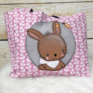 Puede incluir: Bolsa de tela rosa con estampado floral blanco. Un círculo gris con un conejito marrón con un babero rosa de lunares blancos está centrado en la bolsa.