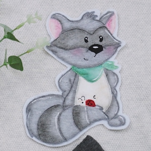 May include: A grey raccoon with a green scarf and a red ladybug on its belly. The raccoon is sitting on a white background.