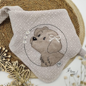 Puede incluir: Una babero de muselina gris y blanco para bebé con un diseño de perro bordado en marrón. El perro tiene un lazo blanco en la oreja y está rodeado de flores blancas bordadas. El texto "Bine naht" está bordado en la esquina inferior derecha.
