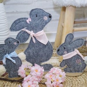 May include: Three gray felt bunny rabbit figurines with pink and blue ribbon accents. The bunnies are standing on a wooden surface with pink flowers in the foreground.