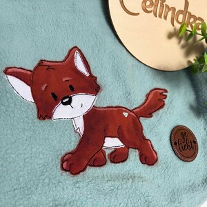 Könnte beinhalten: Eine hellblaue Fleece-Decke mit einem roten Fuchs-Applique und einem braunen Leder-Etikett mit der Aufschrift "ge liebt".