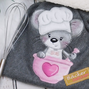 Könnte beinhalten: Grauer Stoffbeutel mit Kordelzug mit einer niedlichen Comic-Maus, die einen Kochhut trägt und einen Schneebesen hält. Die Maus befindet sich in einer rosa Schüssel mit einem Herzen. Der Beutel hat ein gelbes Etikett mit der Aufschrift "Bäcker Meister".
