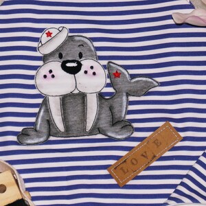 May include: A blue and white striped fabric with a gray and white cartoon walrus wearing a red and white sailor hat. The walrus has a brown leather tag that says "LOVE" on it.