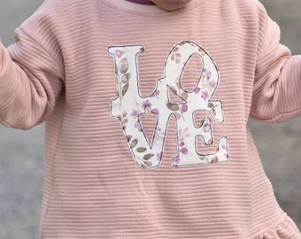 May include: A light pink ribbed-knit dress with a floral "LOVE" applique. The dress features a ruffled hem and long sleeves. A small logo is visible in the upper left corner. The child is wearing a pink headband.