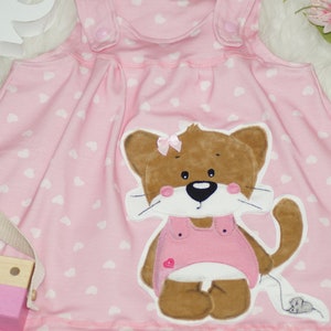 May include: Pink sleeveless dress with a white heart pattern. A brown cat with a pink bow and a pink dress is embroidered on the front of the dress.