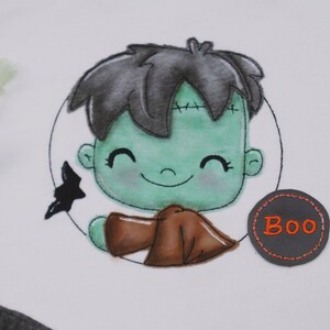 Könnte beinhalten: Ein grünes Monster mit schwarzen Haaren und einem braunen Umhang lächelt. Das Monster befindet sich in einem Kreis mit einer schwarzen Fledermaus und dem Wort "Boo" in einem grauen Kreis.