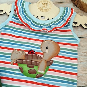 Puede incluir: Una camiseta sin mangas a rayas con un cuello de volantes azul y una tortuga marrón con una mariquita roja en la espalda. La camiseta tiene rayas rojas, azules, verdes y rosas.