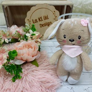 以下が含まれることがあります： ピンクのリボンとピンクのドット柄のスタイをつけたベージュのぬいぐるみウサギ。ウサギは、花輪と「La Kiki Design」と書かれた木製の看板のあるピンクのふわふわの表面に座っています。