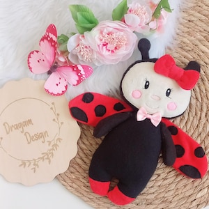 Op de afbeelding: Een pluche lieveheersbeestje speelgoed met zwarte en rode vleugels, een roze strik en een wit gezicht met geborduurde ogen en een glimlach. Het speelgoed zit op een gevlochten mand.