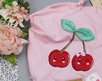 May include: A light pink and white gingham fabric pouch featuring an appliqué of two red cherries with smiling faces, green leaves, and a small pink bow. The pouch is finished with a pink pom-pom trim at the top edge and a small rectangular label.