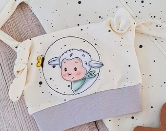 May include: A white baby hat with a gray brim and a cute cartoon lamb with a butterfly design. The hat has a white background with black polka dots.