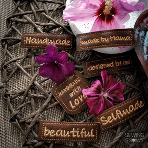 Op de afbeelding: Handgemaakte lederen labels met verschillende zinnen, waaronder "Handgemaakt met liefde", "Zelfgemaakt", "Mooi", "Handgemaakt", "Gemaakt door mama" en "Ontworpen door mij". De labels zijn bruin met gouden stiksels en zijn gerangschikt op een geweven bruine achtergrond.