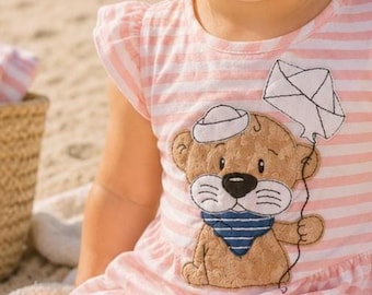 May include: A light pink and white striped short-sleeve dress featuring a brown bear appliqué wearing a white sailor hat and a blue and white striped neckerchief, while holding a white kite on a string against a soft, neutral-toned background.