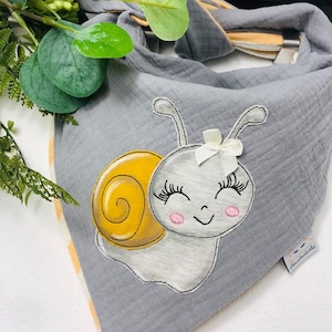 Puede incluir: Un babero de tela gris y amarillo para bebé con un lindo diseño de caracol bordado. El caracol tiene una concha amarilla, mejillas rosadas y pestañas. El babero tiene un lazo blanco en la parte superior de la cabeza del caracol. El babero tiene un borde a rayas de color naranja claro y blanco.