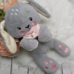 Puede incluir: Un conejito de peluche gris con orejas largas, detalles rosas en las orejas y las patas, y una bufanda rosa y blanca alrededor del cuello.