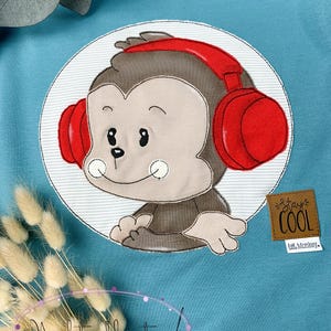 May include: A blue t-shirt with a white circle containing a brown monkey wearing red headphones. The monkey is smiling and has a small white stripe on its chest. The text "Stay Cool" is on a brown tag with the text "lil' Monkey" below it.