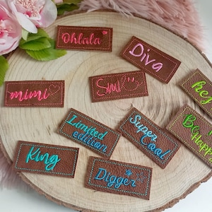 Könnte beinhalten: Zehn braune Leder-Etiketten mit bunten gestickten Texten. Die Etiketten beinhalten die Wörter "Oh Lala", "Diva", "Smile", "Mimi", "Limited Edition", "Super Cool", "Hej!", "Be Happy", "King" und "Digger".