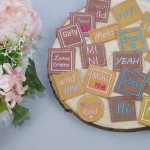 以下が含まれることがあります： 様々なフレーズとデザインが施された、小さな正方形の革のパッチの集まり。 「Lama Drama」、「Wild One」、「Mini Me」、「Hey」、「No」、「Star」、「Yeah」、「Mott」、「Girly」、「Frechi Id」、「Papar Held」、「Mini Me」、「Ahm Nein」、「BILA BILA BLA」、「Kind」、「Yay!」など。 パッチは木製の表面に配置されています。