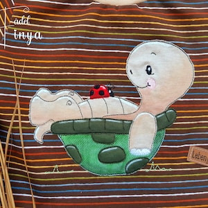 Puede incluir: Una camisa a rayas marrones y blancas con una imagen bordada de una tortuga con una mariquita en la espalda. La tortuga es verde y tiene un caparazón blanco. La camisa tiene el texto "Leben! Lieber lachen" en una etiqueta marrón.