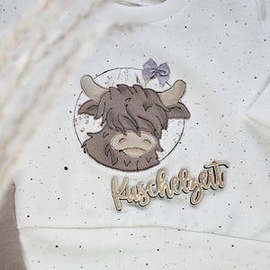 Könnte beinhalten: Weißer Pullover mit einer braunen und weißen gestickten Hochlandkuh mit einer lila Schleife und dem Text "Kuschelzeit" in Gold.