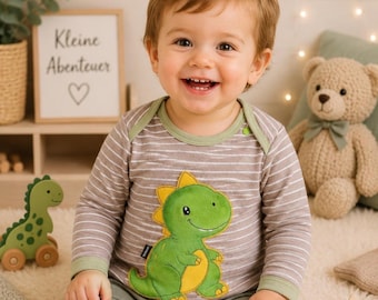 May include: A long-sleeved shirt with a brown and white horizontal stripe pattern, featuring a large green and yellow dinosaur graphic on the front. The shirt has a solid light green trim around the crew neckline and the sleeve cuffs.