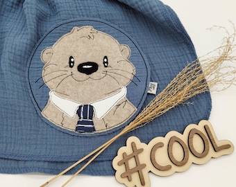 May include: A blue fabric item with a circular patch featuring a cartoon otter wearing a white collared shirt and a blue striped tie. A wooden sign with the word "#COOL" is also visible.