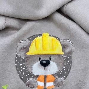 May include: A brown teddy bear wearing a yellow hard hat and an orange vest. The bear is in a circle with white polka dots on a brown background.