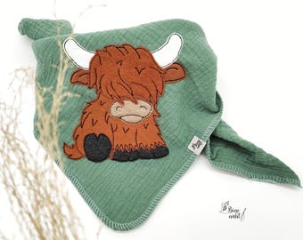 May include: A green muslin baby blanket with a brown and white embroidered Highland cow design. The blanket is made by "Bine Naht" and has a fringed edge.