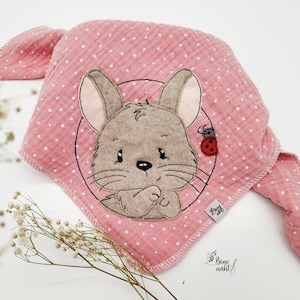 Puede incluir: Babero de bebé de muselina rosa con lunares blancos y un rostro de conejo de dibujos animados bordado en el centro. Una pequeña mariquita roja está al lado del conejo. El babero tiene forma cuadrada con esquinas redondeadas y una textura suave.