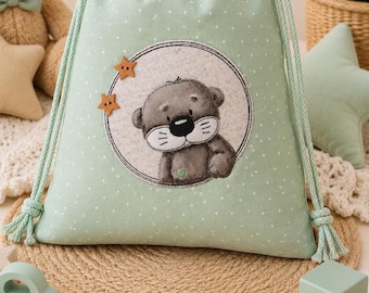 May include: A light mint green drawstring bag featuring a white polka dot pattern and a circular patch with a gray cartoon otter illustration. The bag includes two light green braided rope cords and two small wooden star-shaped buttons attached.