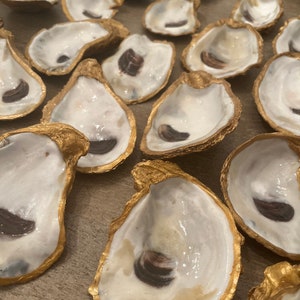 May include: A collection of oyster shells with gold painted edges. The shells are arranged in a random pattern on a wooden surface. Some of the shells have a dark brown interior.
