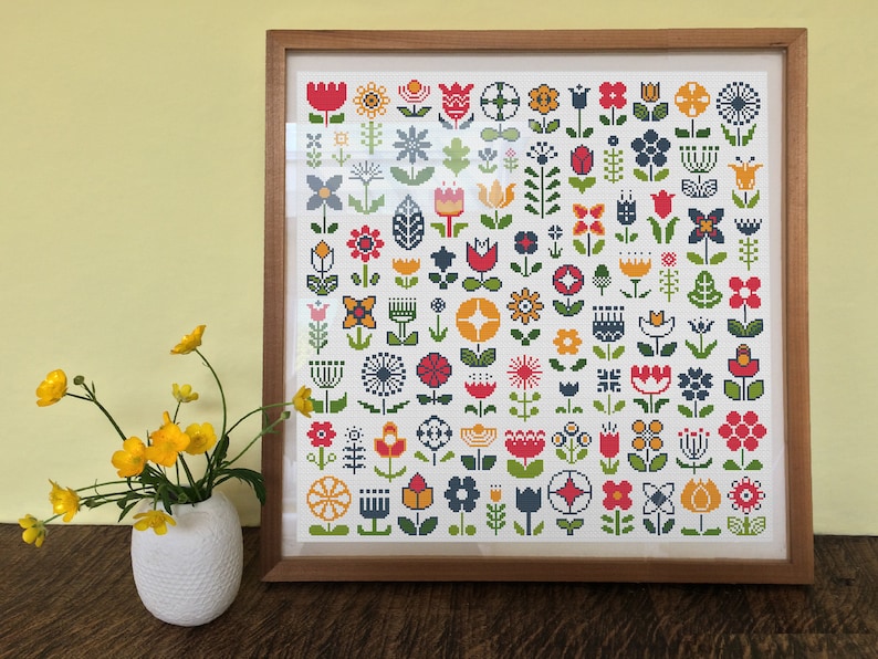 May include: A framed cross-stitch pattern featuring a variety of colorful flowers in a white background. The flowers are in a variety of styles, including tulips, daisies, and other floral designs.