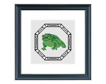 Könnte beinhalten: Ein gerahmtes Kreuzstich-Design mit einem grünen Frosch mit gelben Augen in einem schwarzen und weißen Achteck.