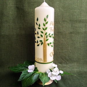 Könnte beinhalten: Weiße Hochzeitskerze mit einem grünen und goldenen Baumdesign und dem Text "Au & Fr" und "2014". Die Kerze ist von weißen Blumen und grünen Blättern umgeben.
