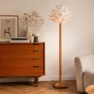 May include: A floor lamp with a tall, slender wooden stand and a square base, topped with a white, multi-layered, petal-like shade that radiates light. The lamp features a clean, vertical design that supports the intricate, tiered light fixture.