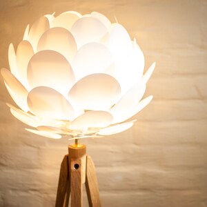 May include: A white floor lamp with a unique, flower-shaped shade made of overlapping white petals. The lamp has a wooden tripod base.