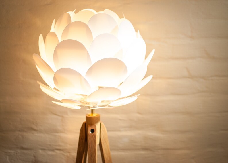 May include: A white floor lamp with a unique, flower-shaped shade made of overlapping white petals. The lamp has a wooden tripod base.