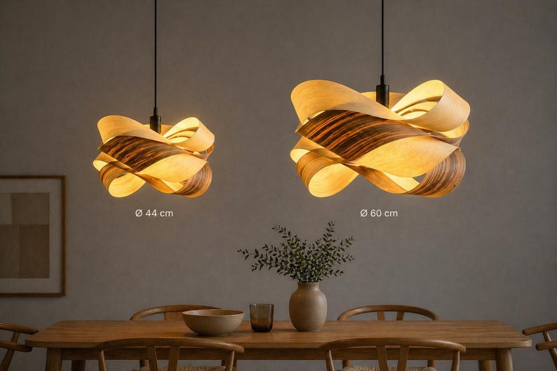 May include: Two pendant lights with a unique design. The lampshades are made of layered wood, with a warm, natural color and a darker, striped pattern. The larger lamp is 60 cm in diameter, and the smaller one is 44 cm in diameter. The lights hang above a wooden dining table.
