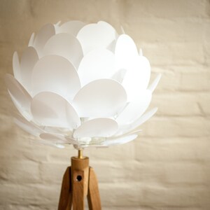 May include: A white, flower-shaped lampshade with a wooden tripod stand. The lampshade is made of many overlapping white petals.