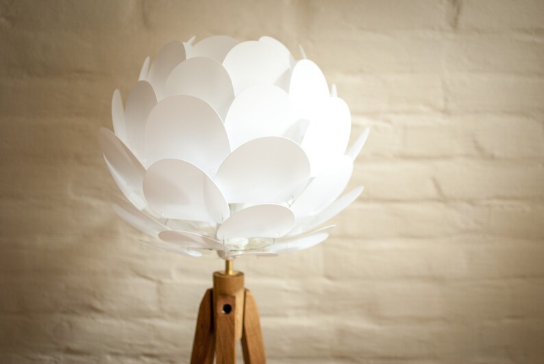 May include: A white, flower-shaped lampshade with a wooden tripod stand. The lampshade is made of many overlapping white petals.