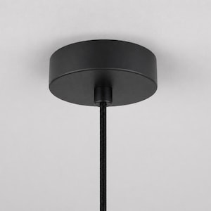 May include: A black ceiling canopy with a cylindrical shape, connected to a black cord. The canopy is mounted against a white ceiling. The cord is a black, braided style. This is a component for a pendant light fixture.