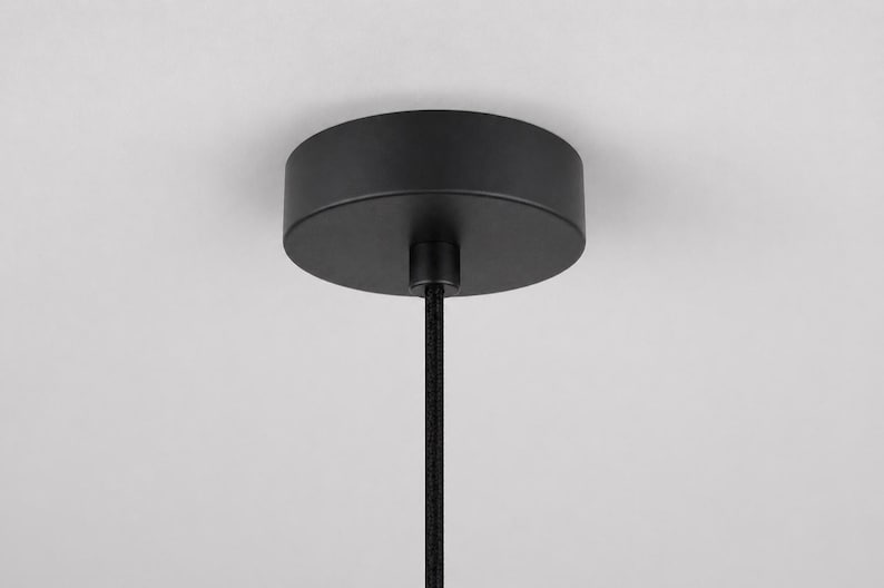 May include: A black ceiling canopy with a cylindrical shape, connected to a black cord. The canopy is mounted against a white ceiling. The cord is a black, braided style. This is a component for a pendant light fixture.