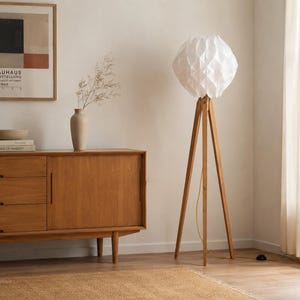Könnte beinhalten: Eine Stehlampe mit einem dreibeinigen Stativfuß aus hellem Holz und einem weißen, geometrisch gefalteten Lampenschirm aus papierartigem Material. Die Lampe steht auf drei schlanken Beinen und hat ein dünnes Kabel, das zu einem schwarzen Schalter führt.