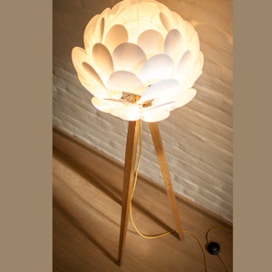 May include: A white floor lamp with a unique flower-shaped shade made of overlapping white petals. The lamp has a wooden tripod base.