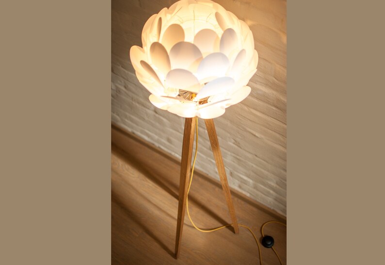 May include: A white floor lamp with a unique flower-shaped shade made of overlapping white petals. The lamp has a wooden tripod base.